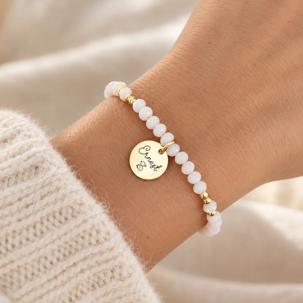Bracelet perles blanches facettées personnalisé avec médaille ronde dorée gravée en acier inoxydable, porté au poignet
